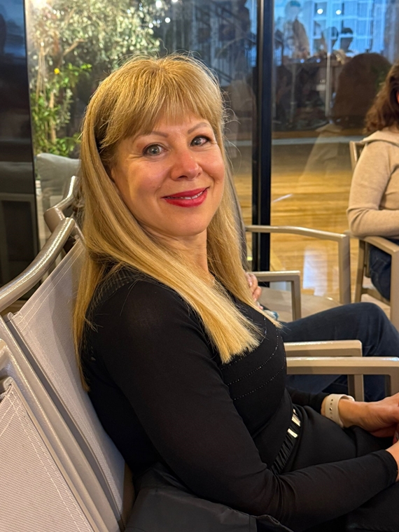 אישה בלונדינית בחולצה שחורה יושבת על כיסא ומחייכת למצלמה, ברקע חלון זכוכית גדול